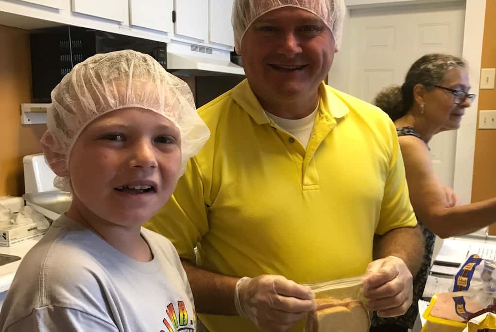 A young boy and adult mentor work assembling sandwiches in a kitchen.