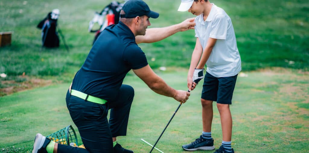 An adult man gives golf lessons to a young boy on a putting green.