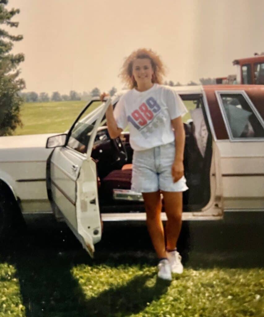 A teen in a 1989 t-shirt and acid-wash shorts stands by the open door of a white station wagon