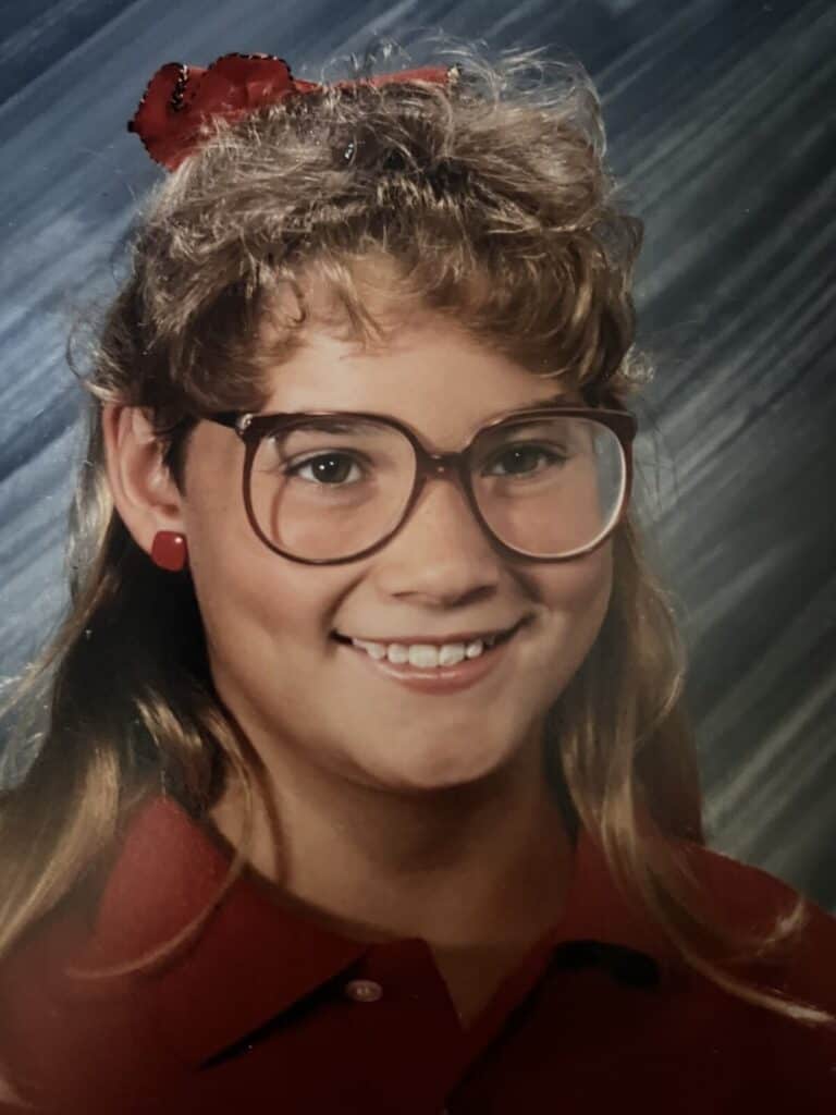 A vintage 80s school photo of a smiling girl with big glasses, feathered bangs and a red bow