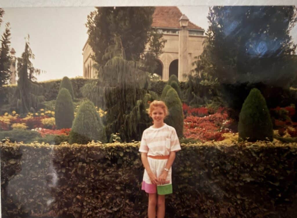 A young girl in pastel clothes poses in front of a formal garden