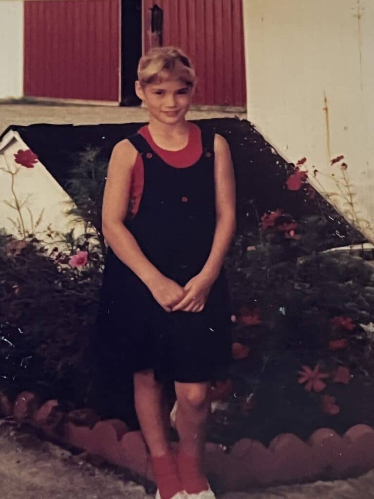 A girl in a black jumper and red shirt stands in front of a flower bed and a red barn