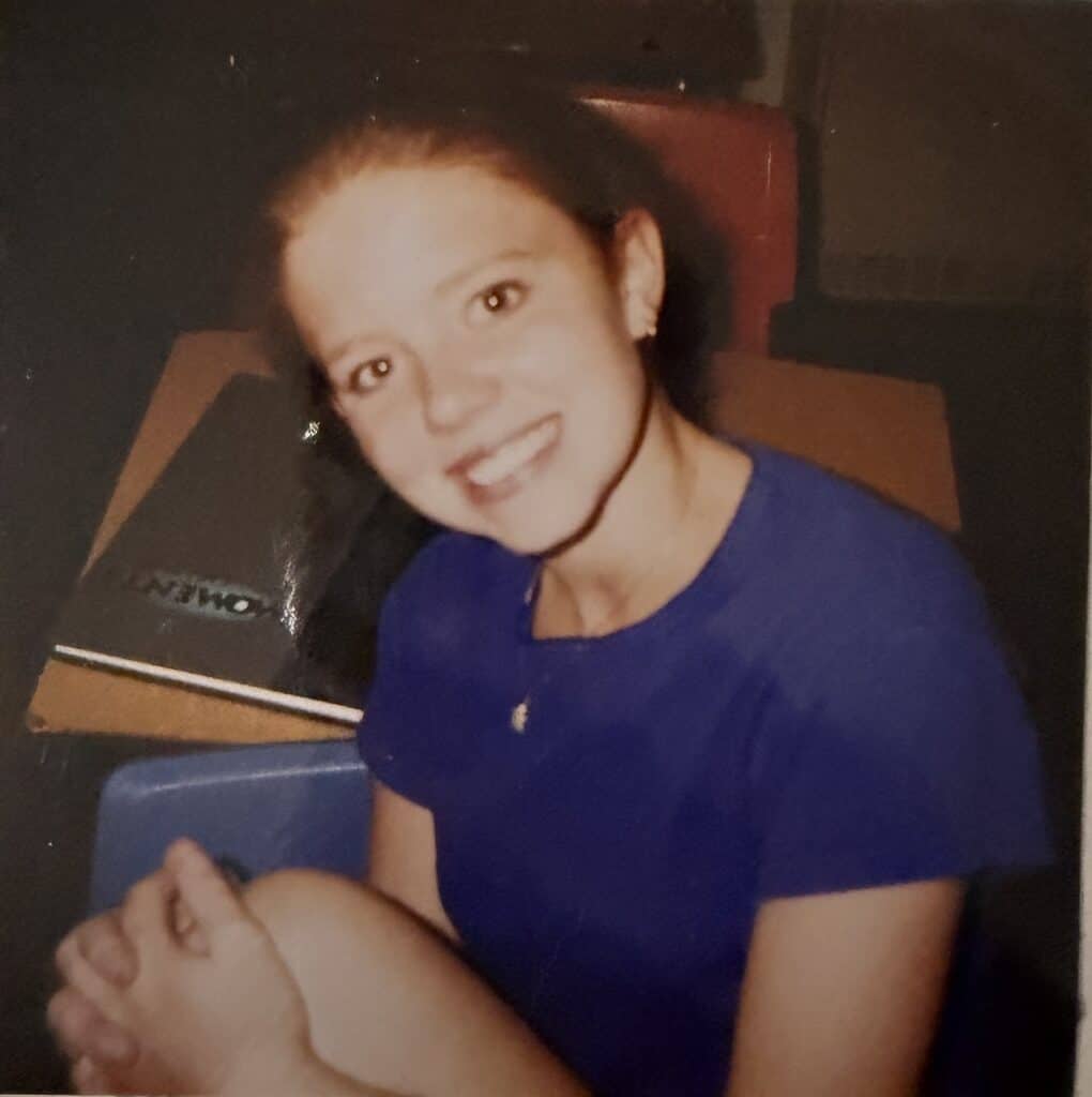 A smiling teen in a purple t-shirt sits next to her school books