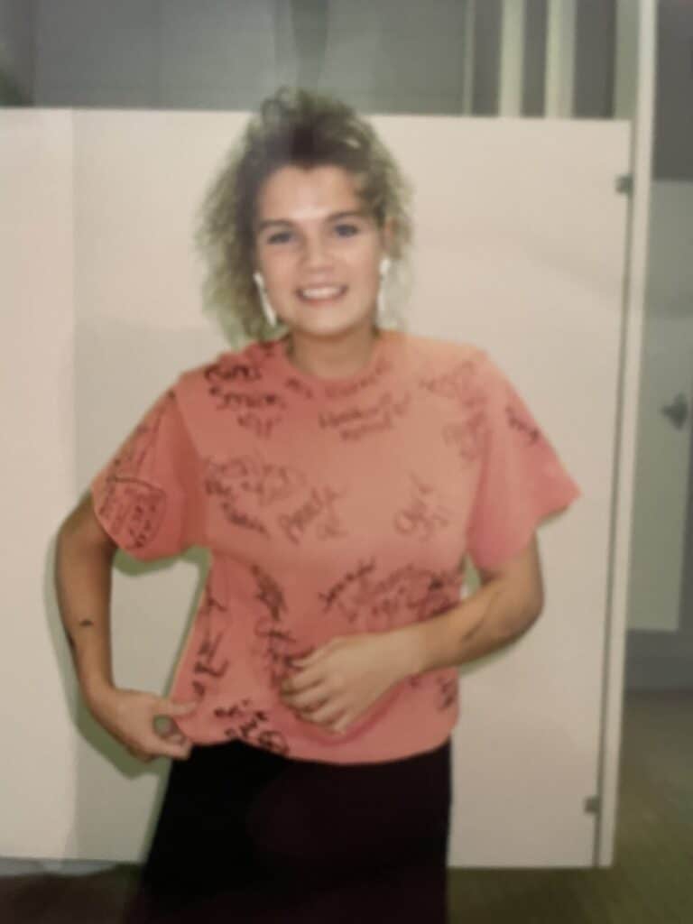 A teen with teased hair wearing a pink t-shirt covered in friends' signatures