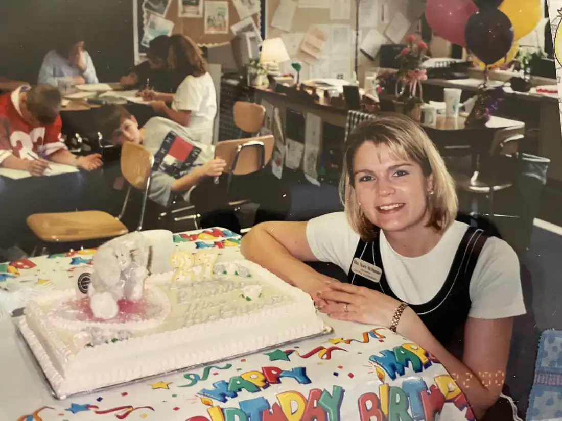 A young woman in a jumper smiles next to a sheet cake and a Happy Birthday tablecloth