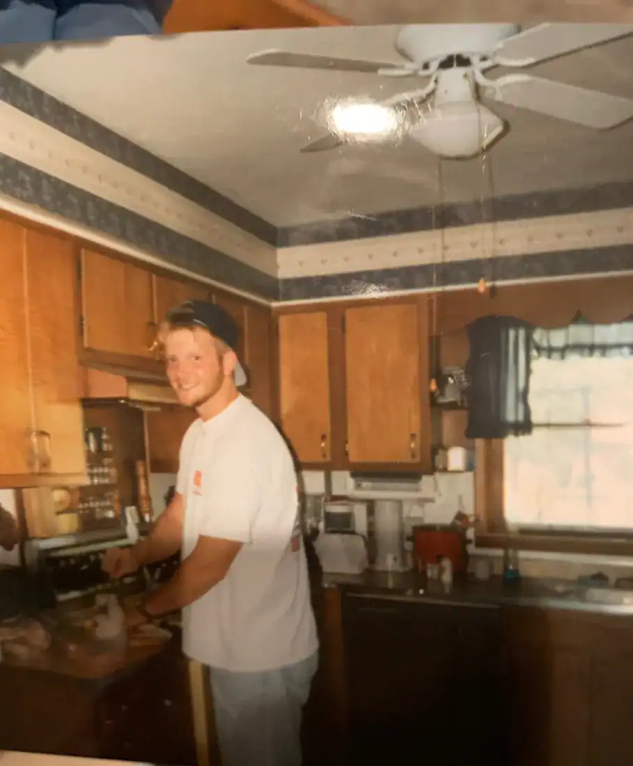A young man in a backwards cap cooking in a wood-cabinet kitchen with a wallpaper border